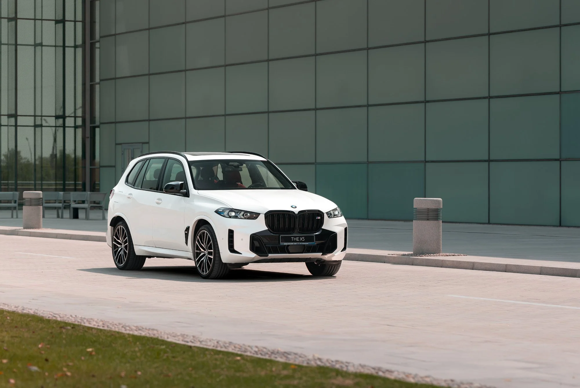 A white BMW X5 M60i SUV parked on a paved area outside a modern building with large glass windows. Photography by Talha Belal.
