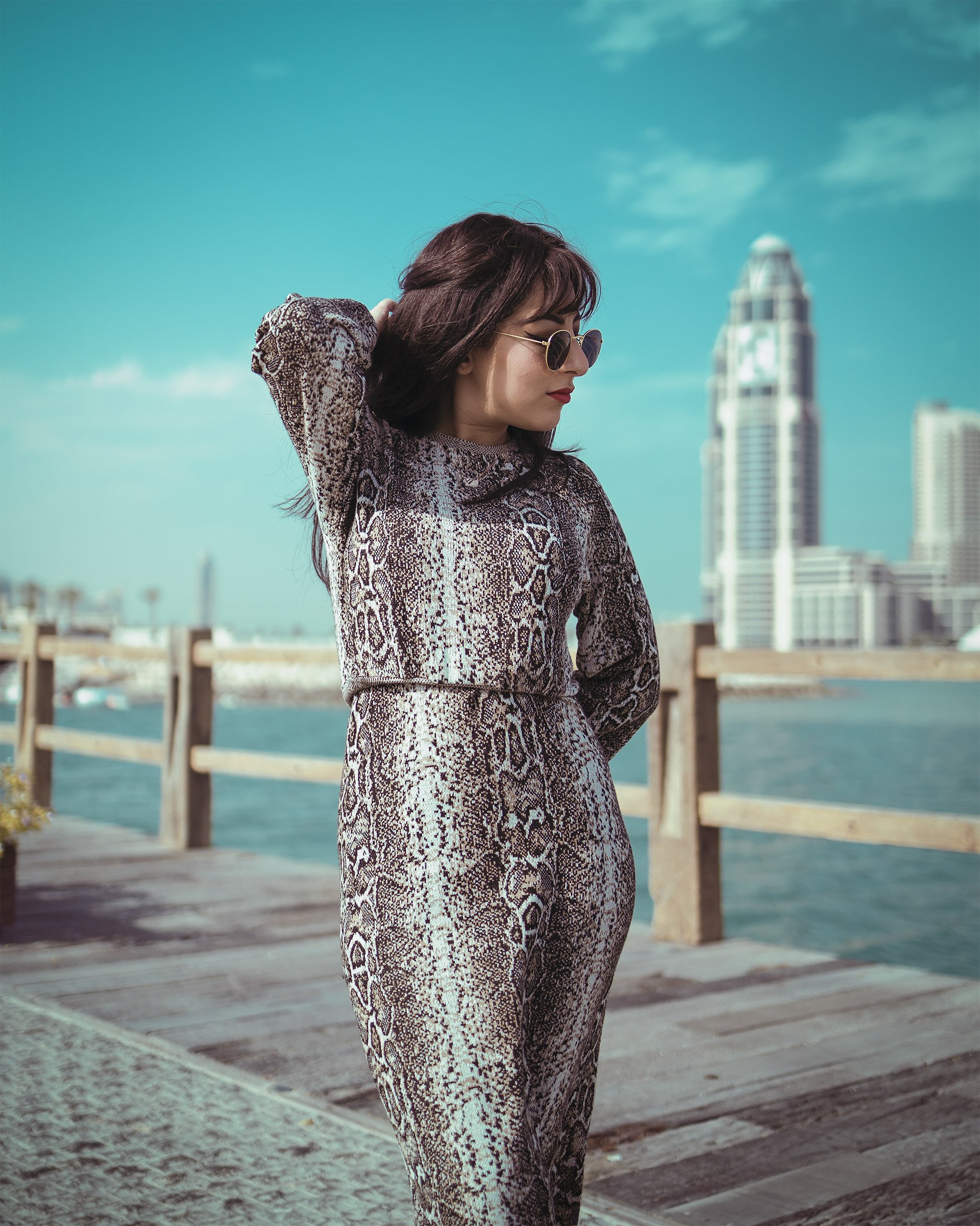 A woman in a snake-print outfit standing on a wooden pier near water with a city skyline and tall buildings in the background.