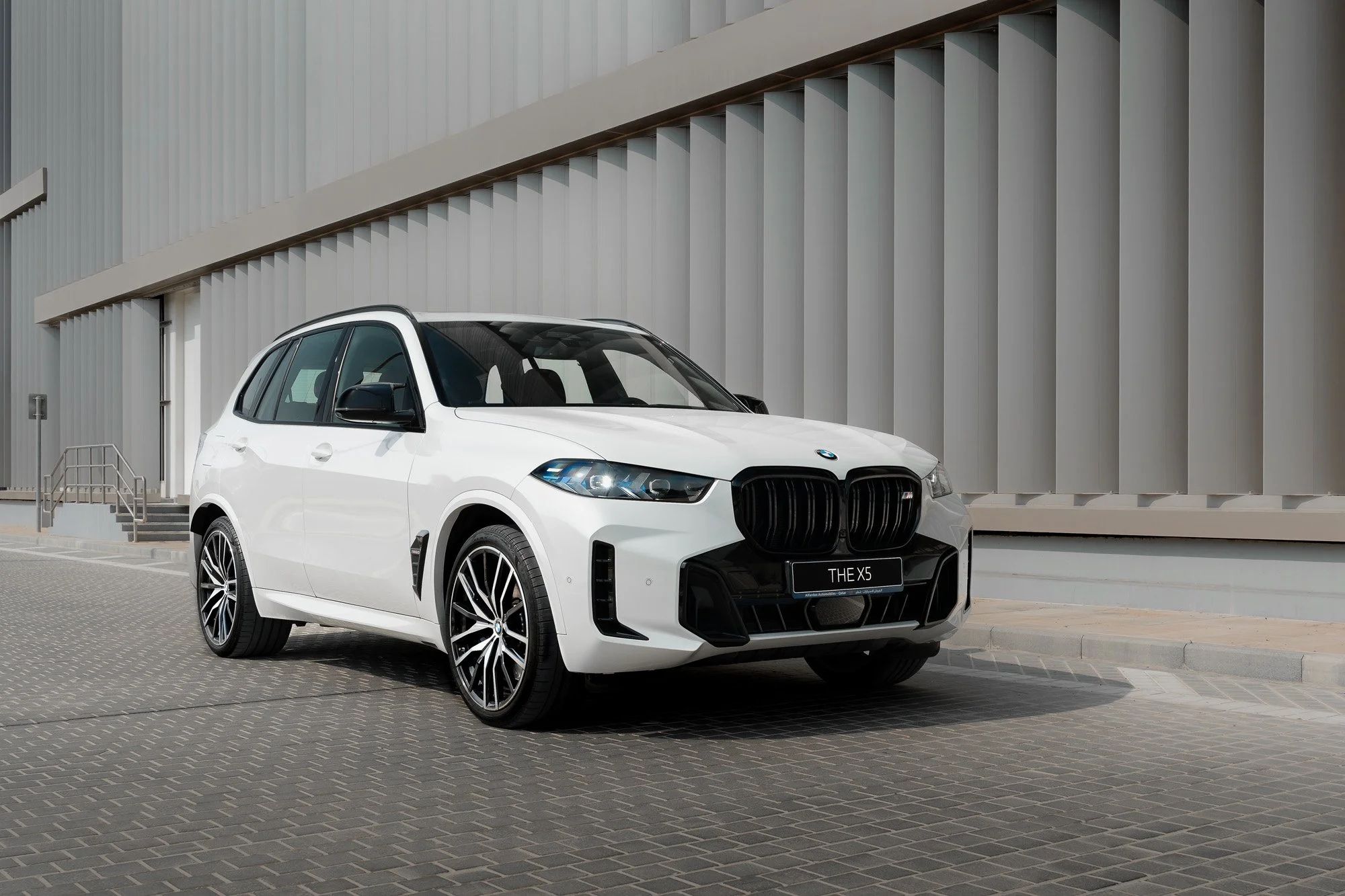 White BMW X5 parked on a brick-paved street in front of a modern gray building with vertical panels.