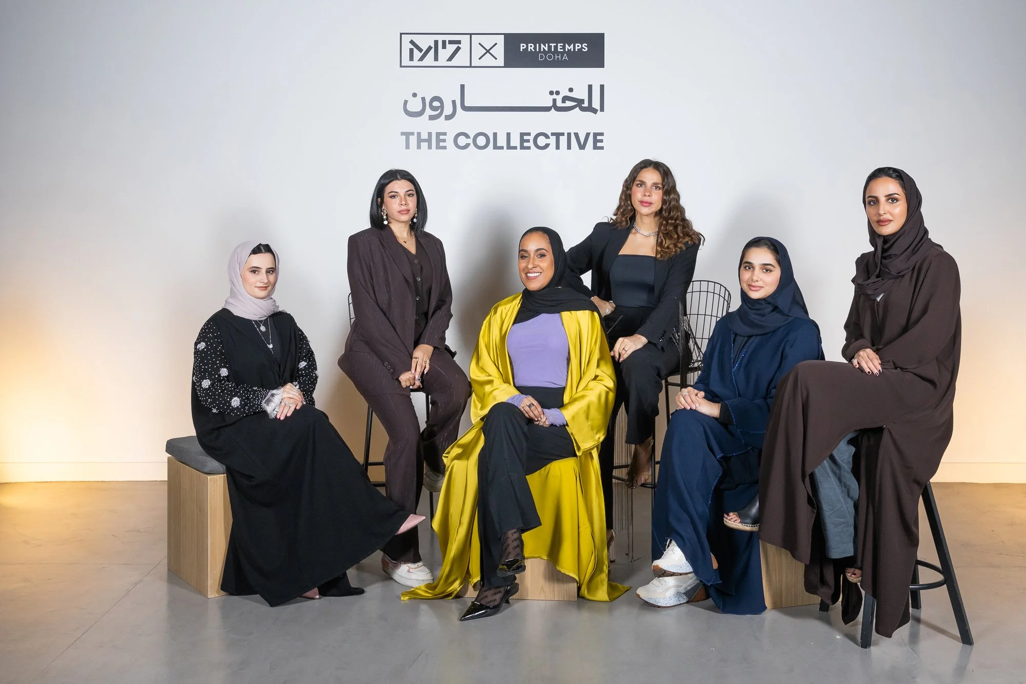 Group of seven diverse women, some wearing hijabs, posing in a modern, minimalist space with white walls for a photo shoot. The backdrop features logos and text reading 'Printemps Doha' and 'The Collective.'
