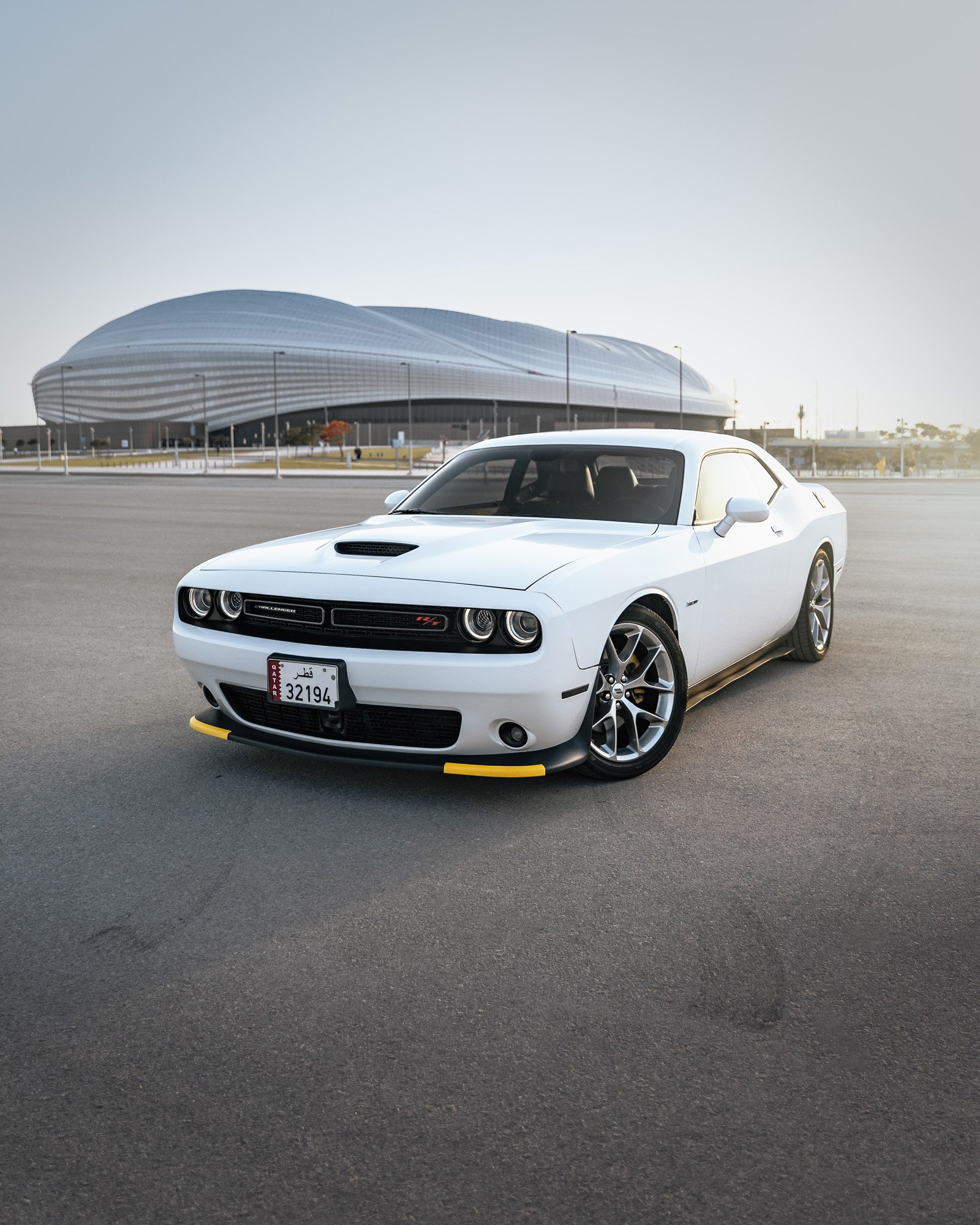 A white Dodge Challenger parked on an empty paved surface with Al Janoub (Al Wakra) Stadium in the background.