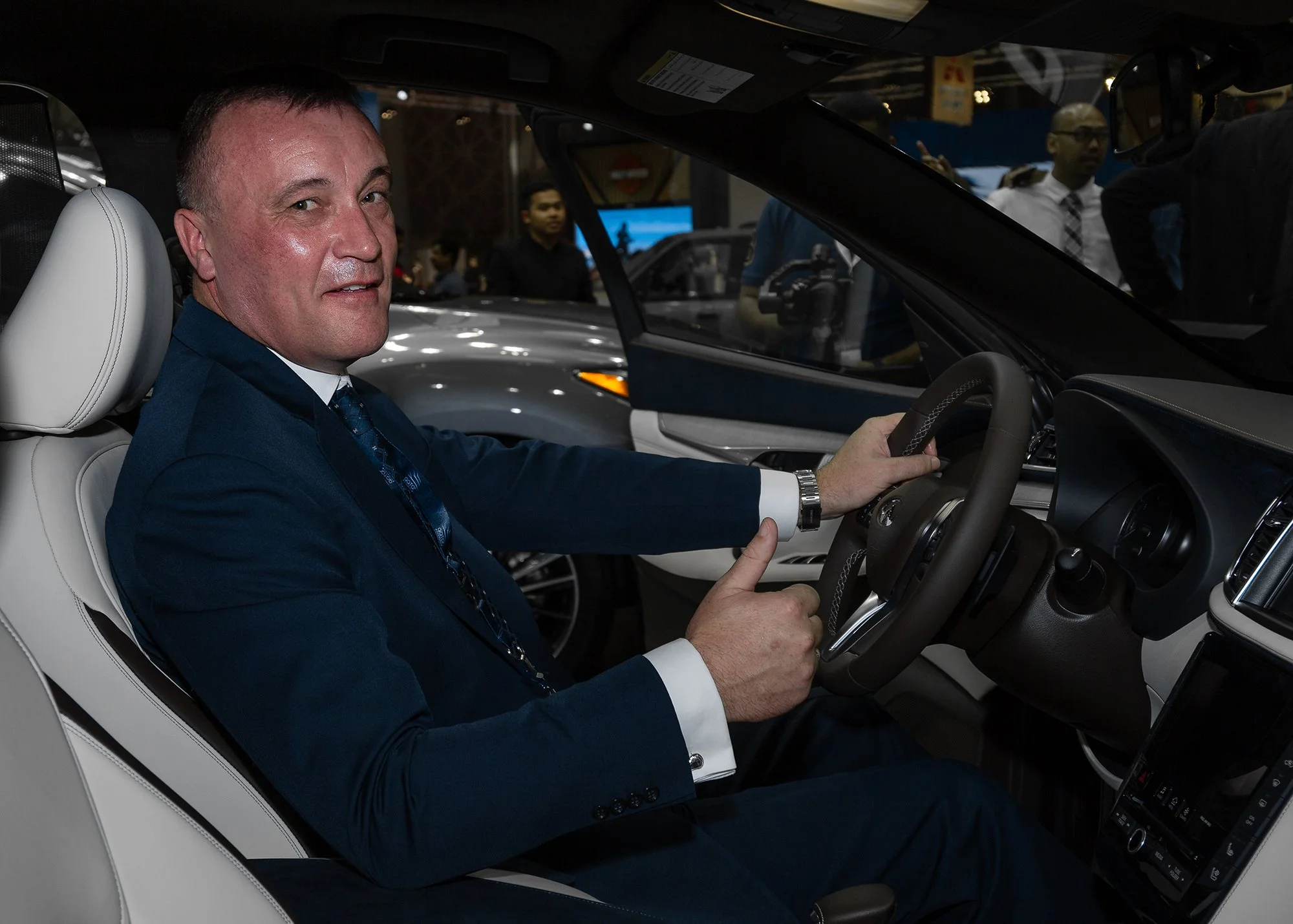 A man in a blue suit and tie sitting in the driver's seat of a car, holding the steering wheel, at an auto show with people in the background.