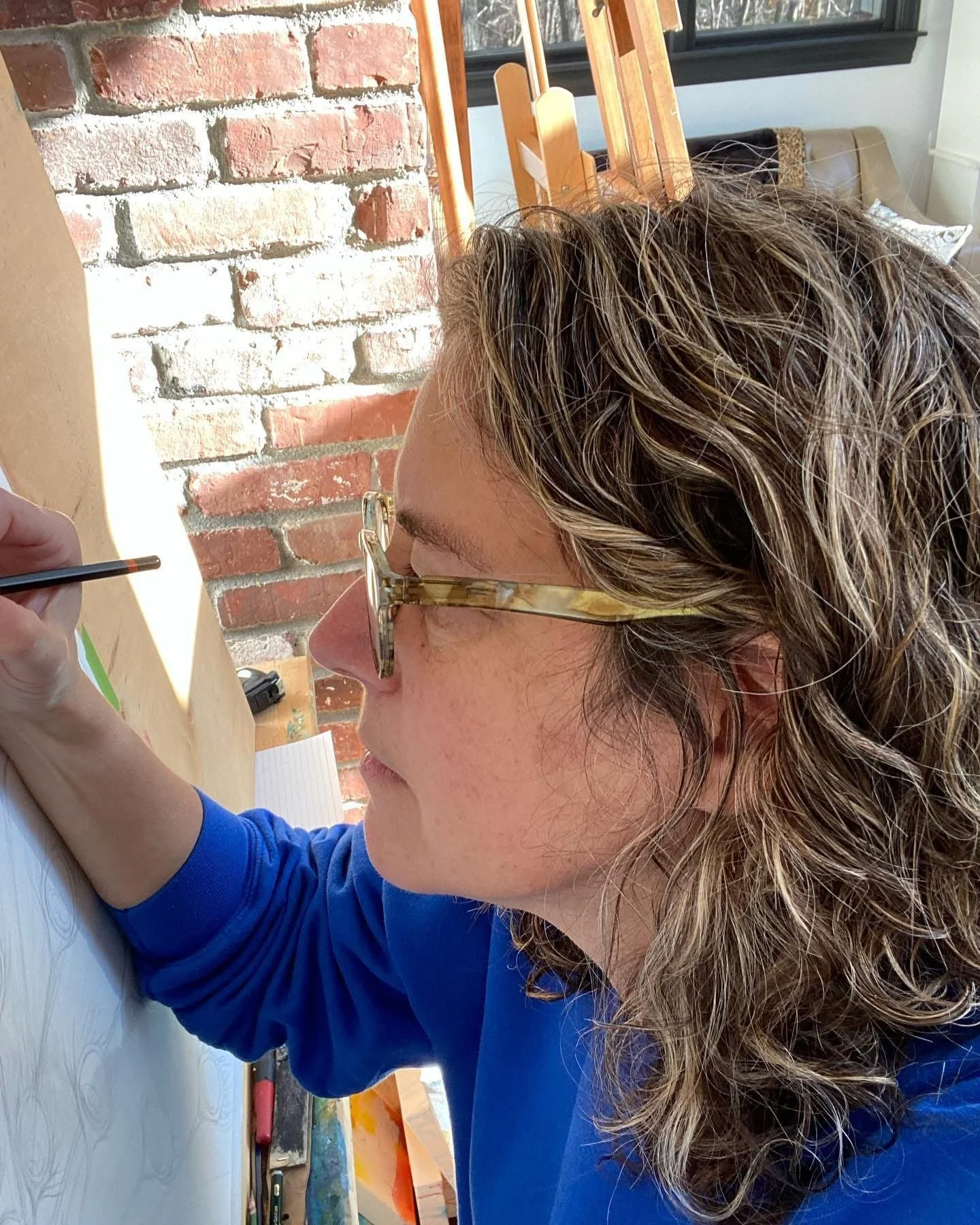 A woman with wavy blonde hair and glasses is painting on a canvas or paper, sitting at an art station in a room with a brick wall and a large window.