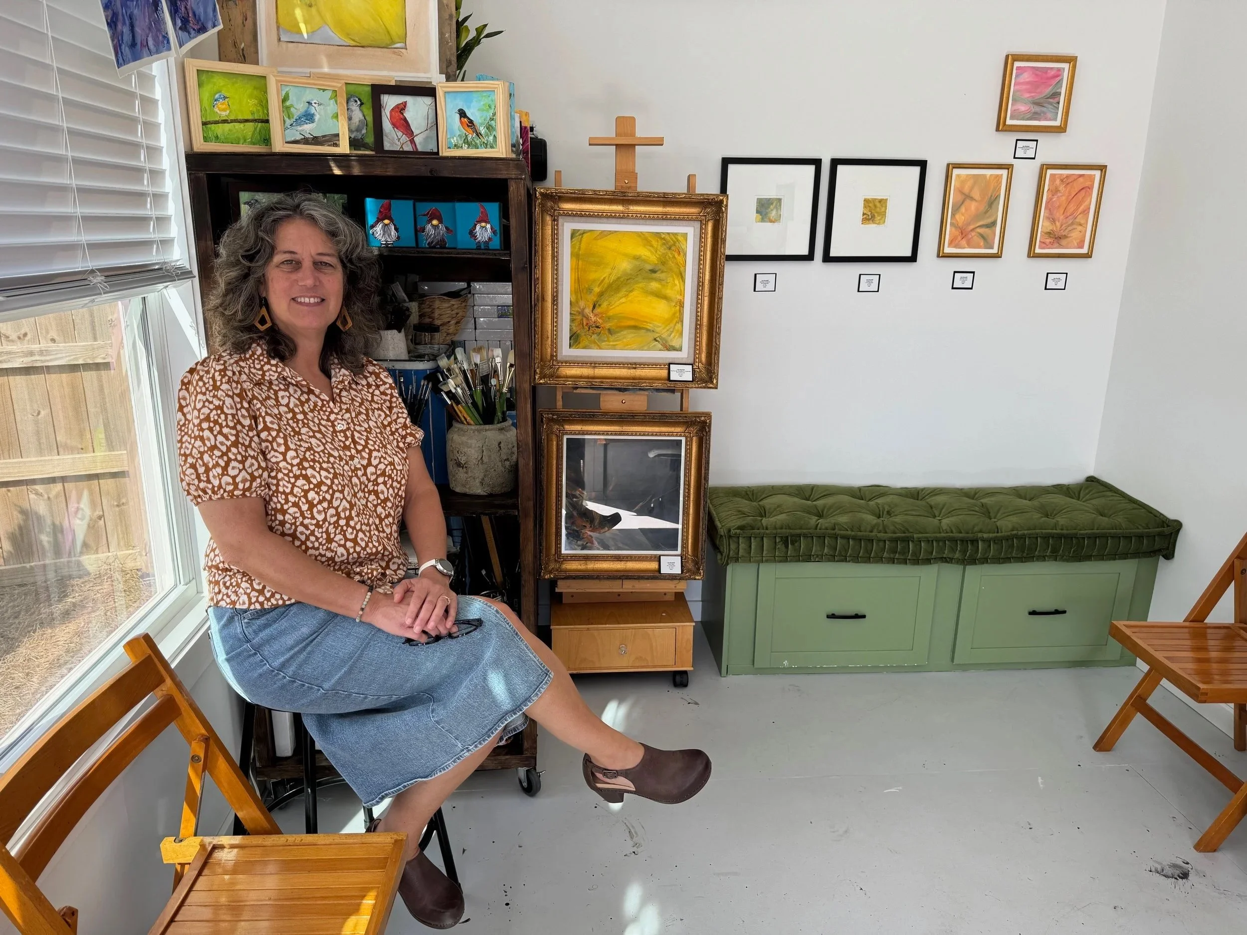 A woman with curly gray hair is sitting on a black chair beside a window with blinds. She is wearing a patterned short-sleeve top, a denim skirt, and brown clogs. Behind her, there is a wooden shelf with framed bird paintings and art supplies. To her right, on the wall, are multiple framed abstract paintings and photographs, and below them a green storage bench with a tufted cushion. There are also wooden folding chairs in the room.
