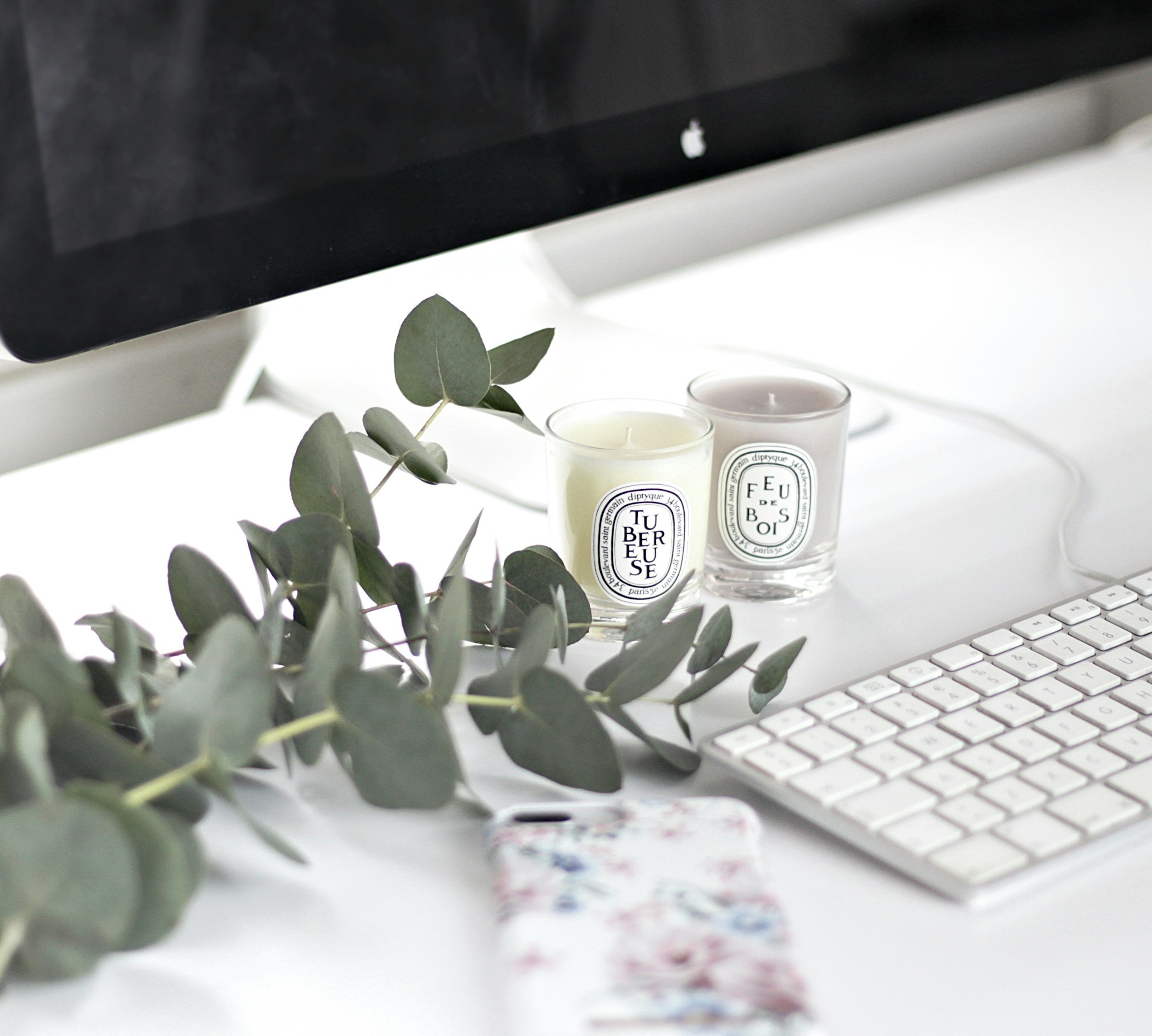 Close-up of a computer monitor with a black frame, a small apple logo on the back, and a white keyboard in front. There are three candles and some eucalyptus leaves placed on the white desk surface.