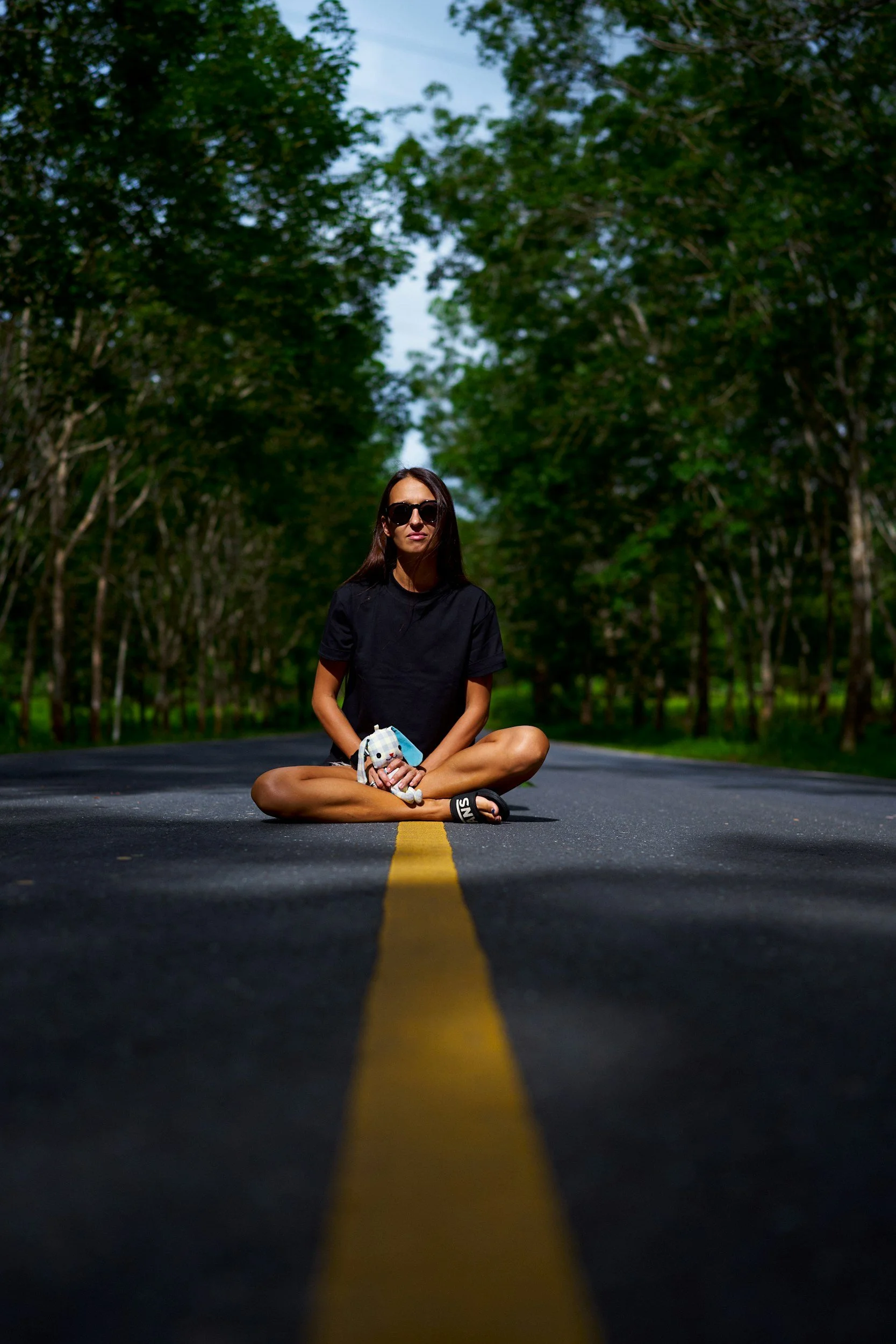 A woman with sunglasses sitting cross-legged in the middle of a road, holding a stuffed toy, surrounded by green trees with a cloudy sky in the background.