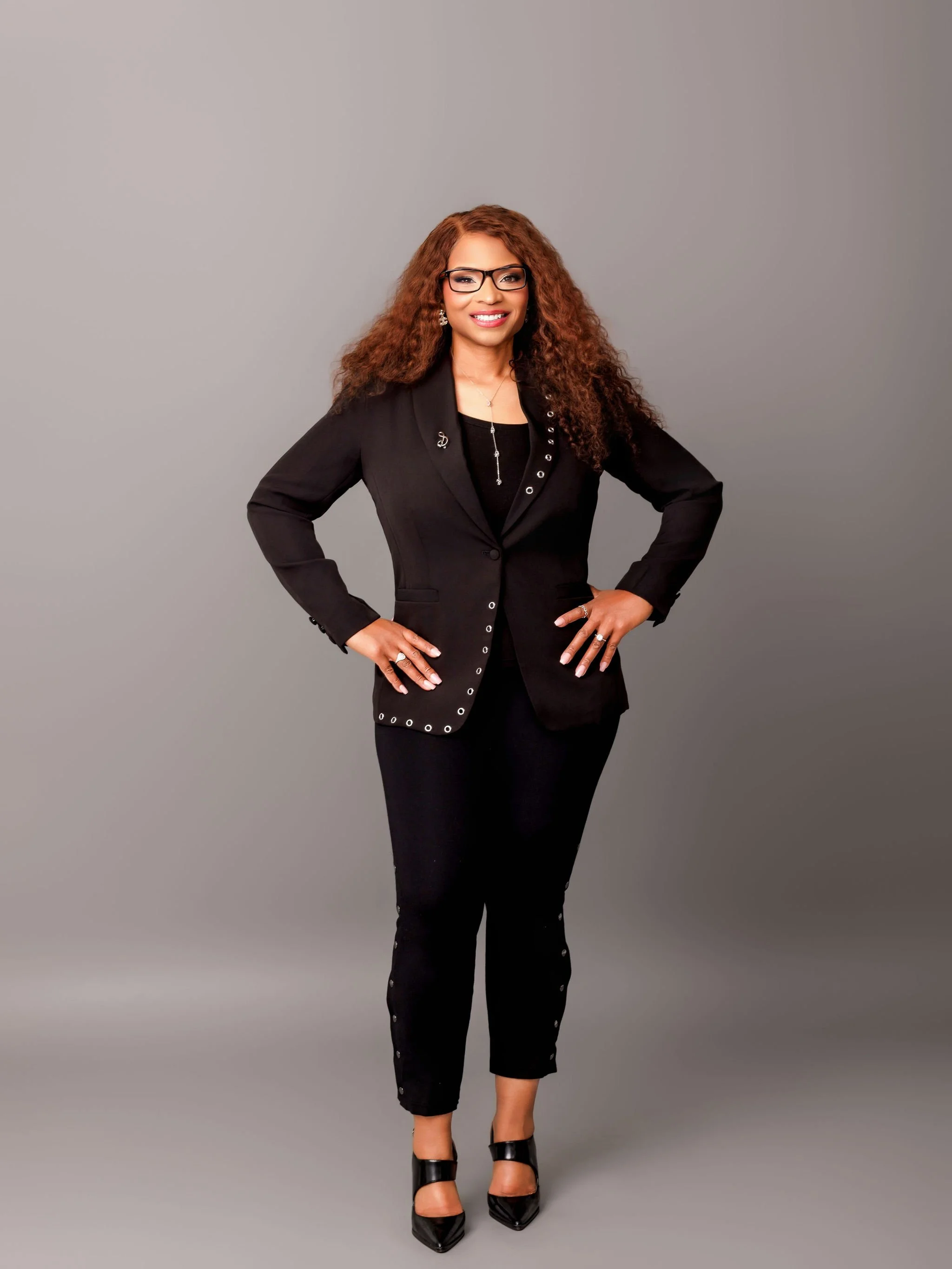A woman with long, curly brown hair and glasses standing against a plain gray background. She is wearing a black blazer with decorative buttons, black pants with buttons down the sides, and black high-heeled shoes. She is smiling with her hands on her hips.