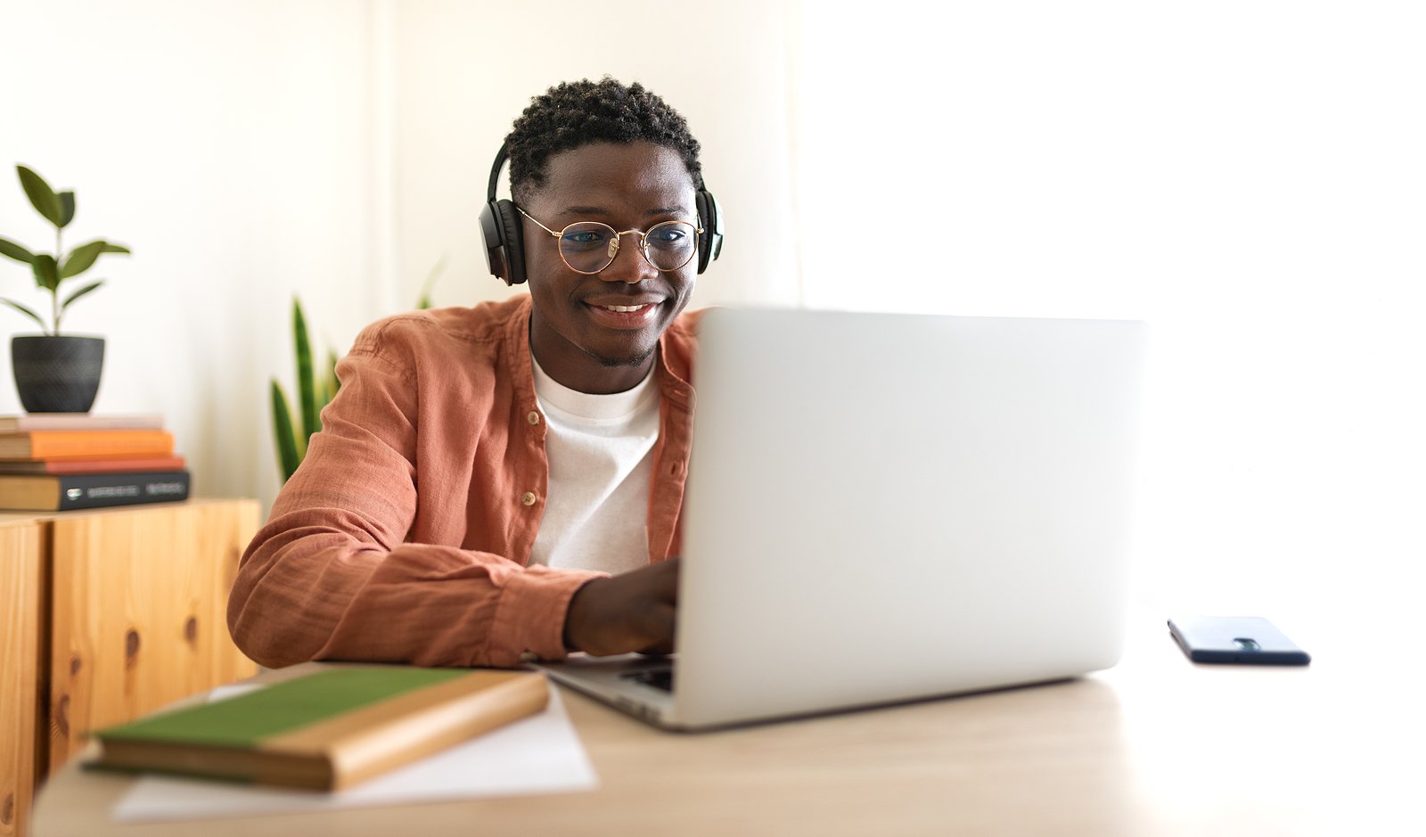 A young man with glasses and headphones working on a laptop at a desk, smiling, with a potted plant and a wooden cabinet in the background.