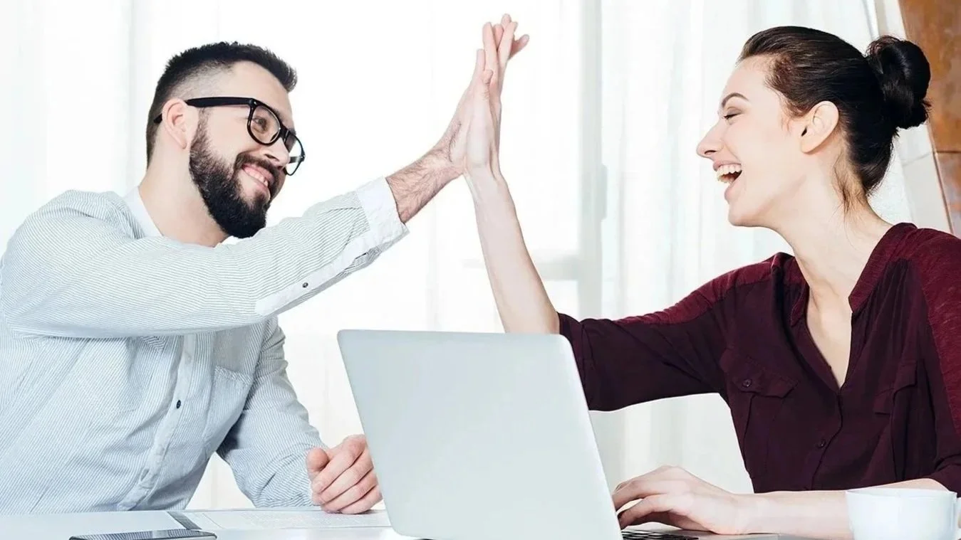 A man and woman giving each other a high five in an office setting, sitting at a desk with a laptop and smartphone, smiling happily.