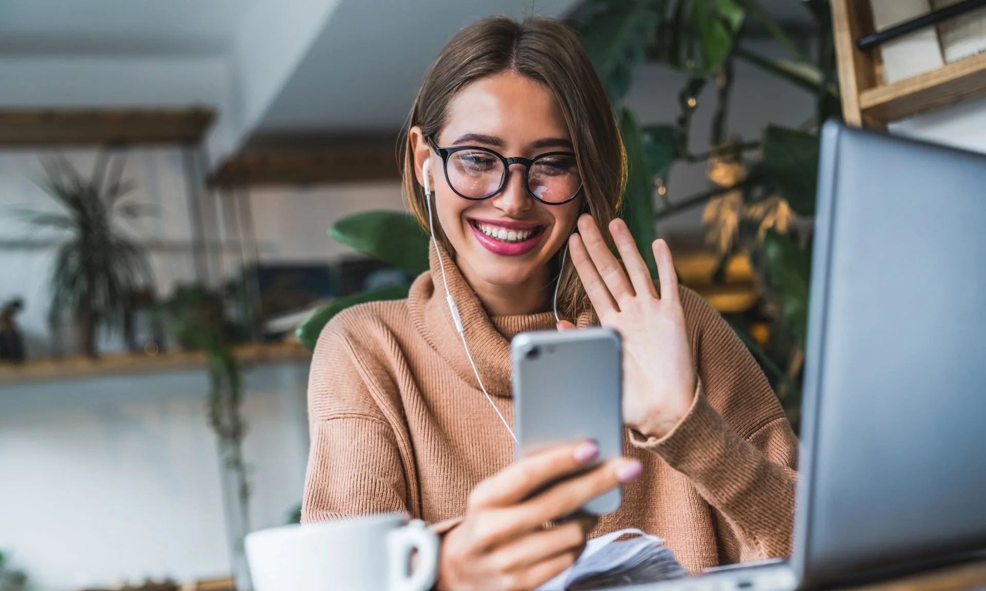A young woman with glasses smiling and waving while looking at her smartphone, sitting in front of a laptop in a cozy indoor setting with green plants.