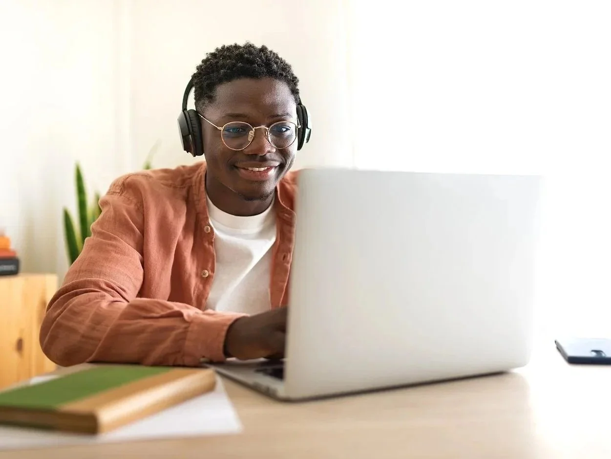 Young man wearing glasses and headphones using a laptop at a desk.