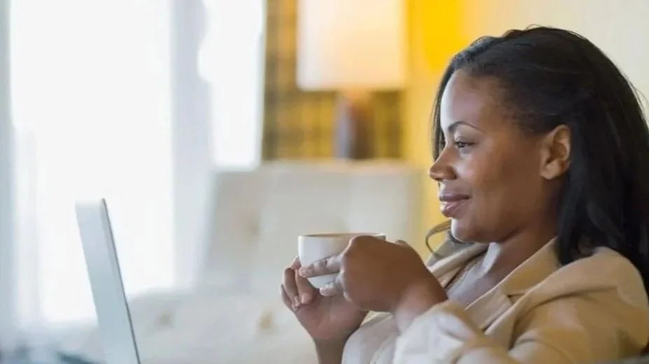 A woman sitting at a table, holding a white mug, looking at a laptop screen with a smile in a well-lit room.