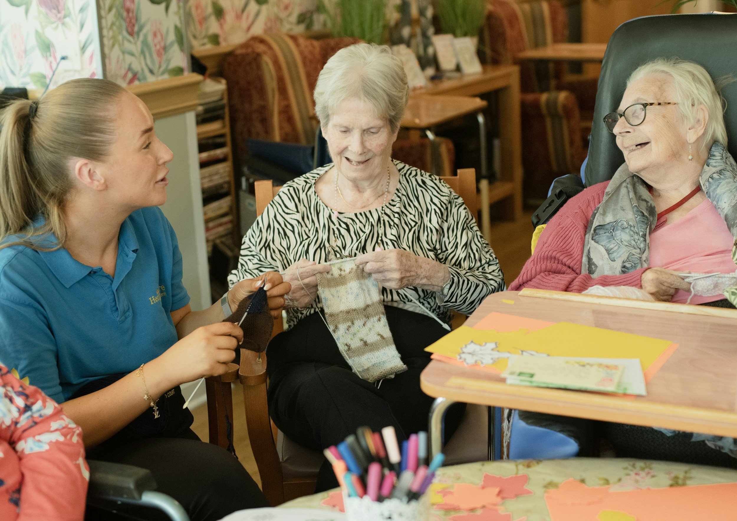 A young woman and two elderly women are sitting at a table, engaging in knitting and craft activities in a cozy, decorated room.