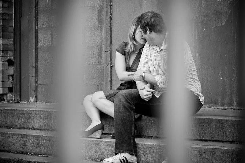 A couple sitting close on a sidewalk, touching foreheads and holding hands through a blurred vertical frame.