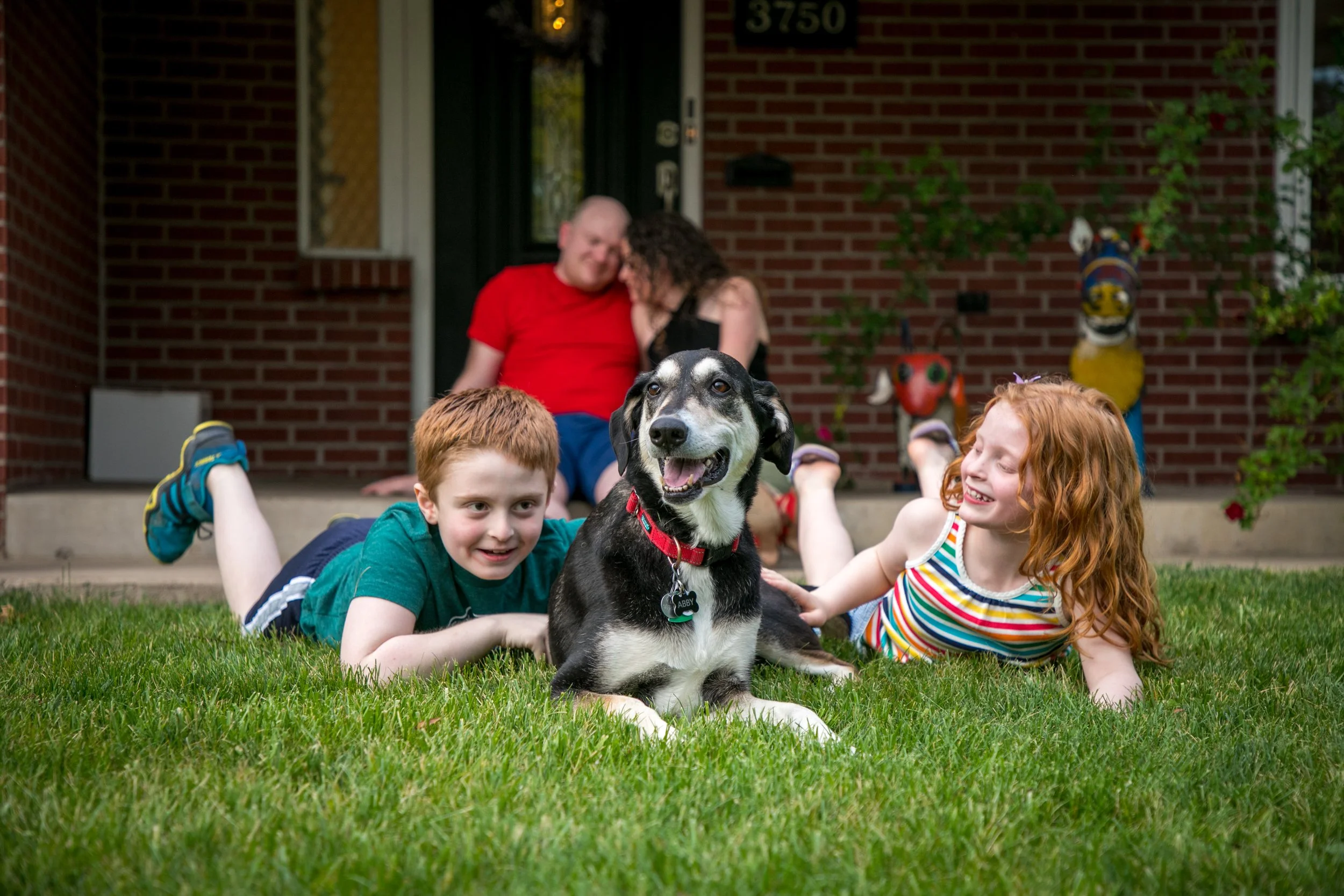 Children and a dog lying on the grass in front of a house with a man and woman sitting on the porch in the background.