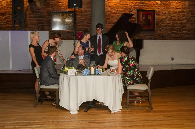 Group of people at a formal dinner, with a woman in a white dress and a man in a suit seated at a round table, others standing and talking in a room with brick walls and a grand piano.