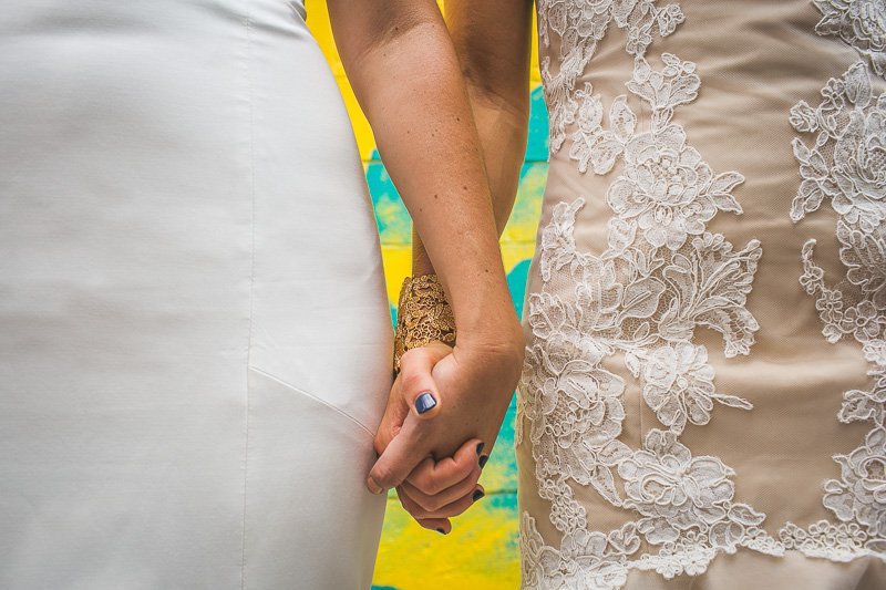 Two women holding hands, one wearing a white dress and the other wearing a beige lace dress, outdoors with a colorful background.