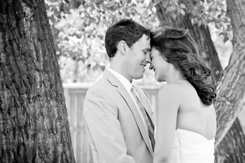 A black-and-white photo of a couple with foreheads touching, smiling, outdoors between trees.