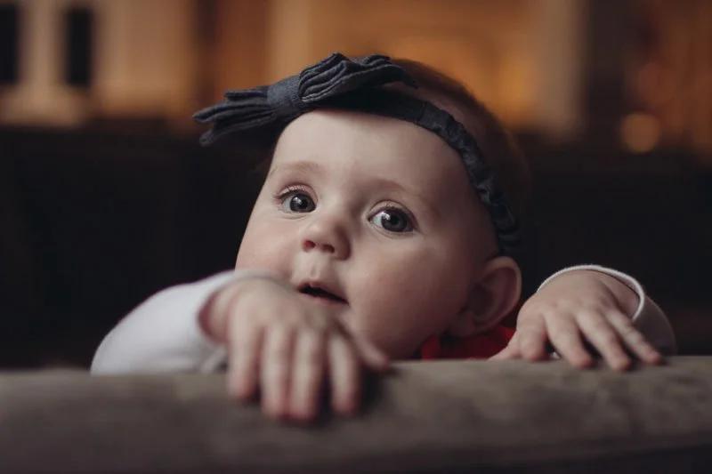 A baby girl with a bow headband reaching over a surface with wide eyes.