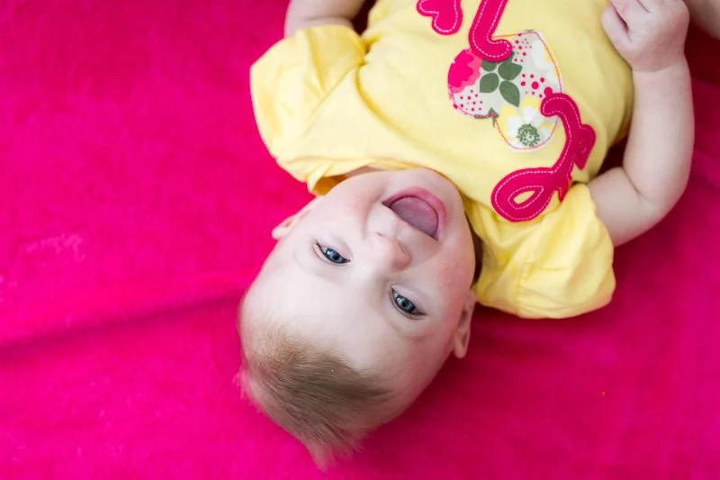 A baby lying on a pink surface, smiling and looking at the camera, wearing a yellow shirt with colorful designs.