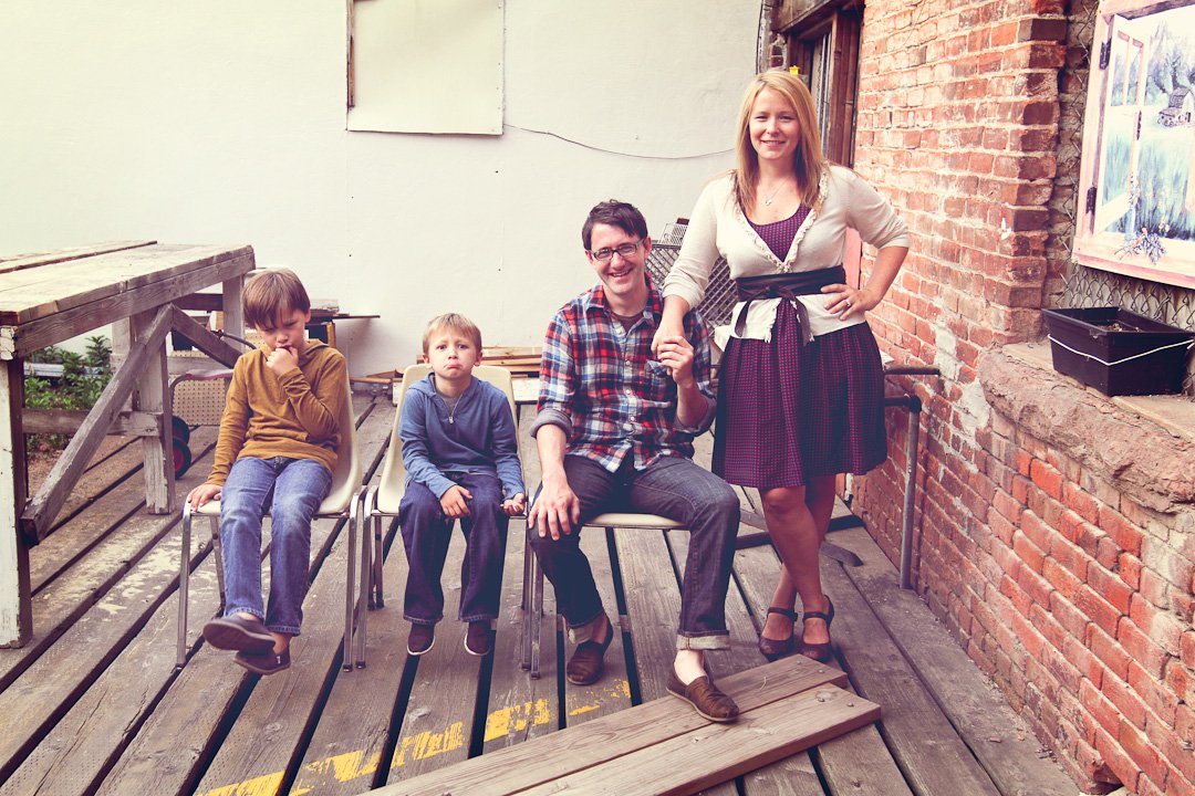 Family of four on a wooden deck outside, with brick and white walls in the background. The adults are sitting and standing, holding hands, while two children sit on chairs.