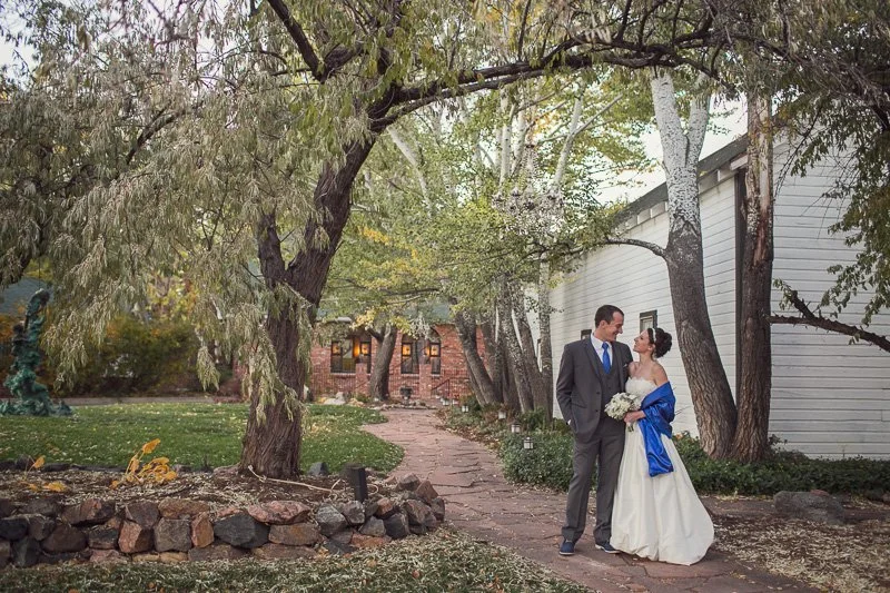 A bride and groom standing on a stone pathway under trees, gazing at each other, with a house in the background