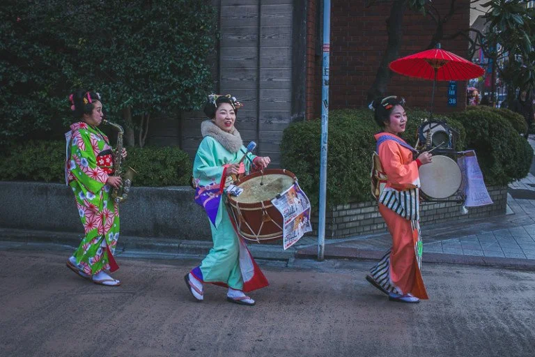 Three women in colorful traditional Japanese kimonos walking on a street, with one holding a musical instrument, another carrying a drum, and the third holding a parasol and drum, during a cultural parade or festival.