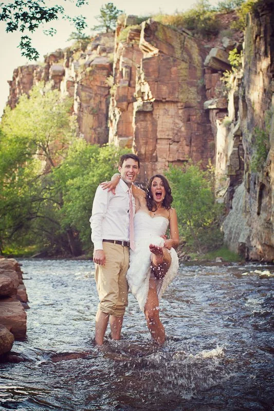 A happy couple in wedding attire standing in a river with a rocky cliff and green trees in the background.