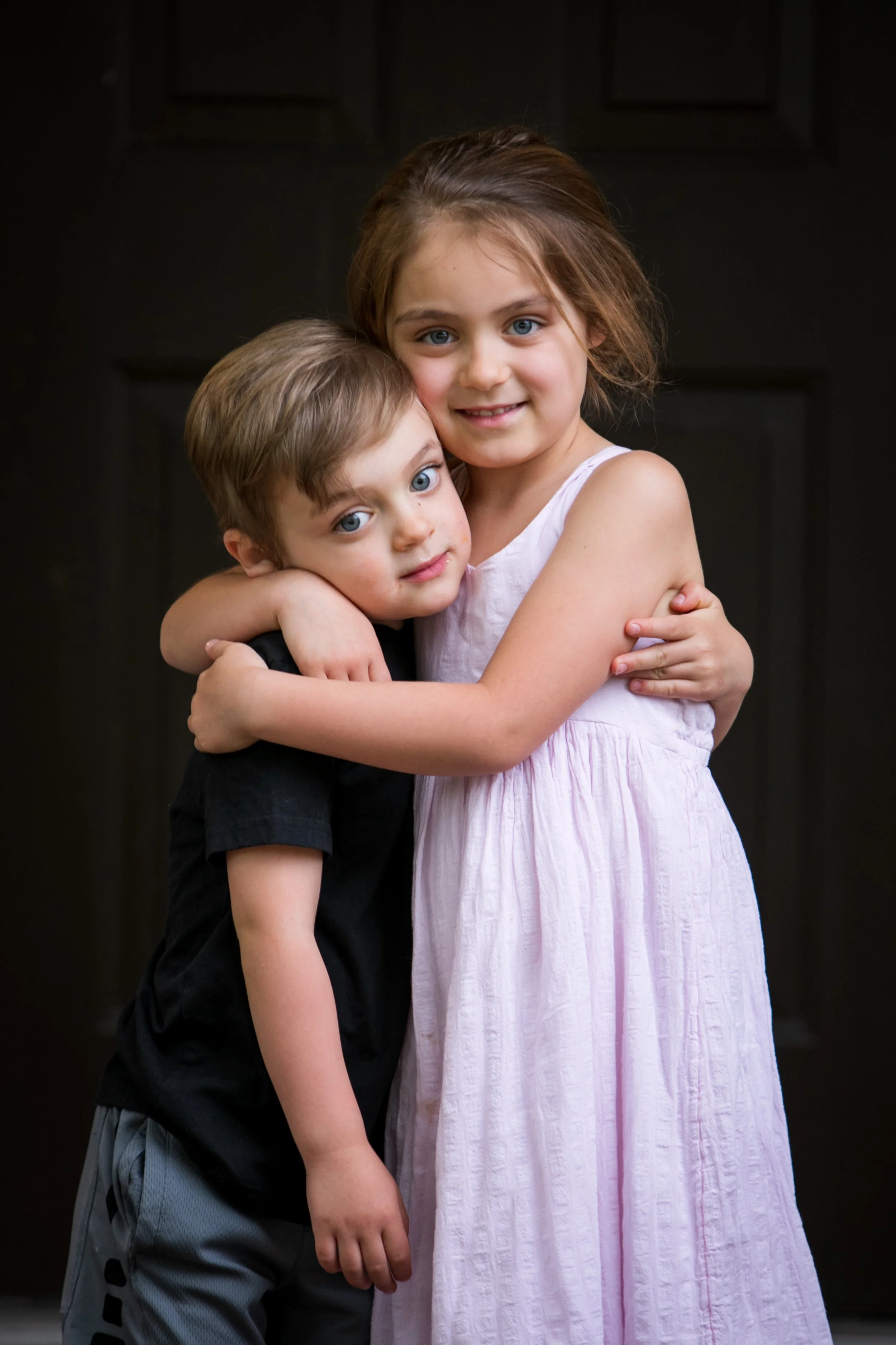 A young girl in a pink dress hugging a young boy in a black shirt, both with blue eyes, standing in front of a dark background.