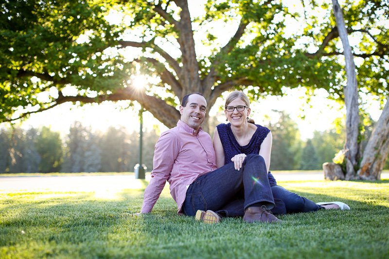 A smiling man and woman sitting on grass in a park with a large tree and sunlight in the background.