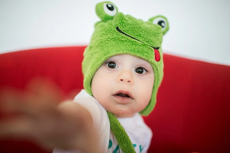Baby wearing a green frog hat with large eyes, tongue, and smile, sitting against a red background.