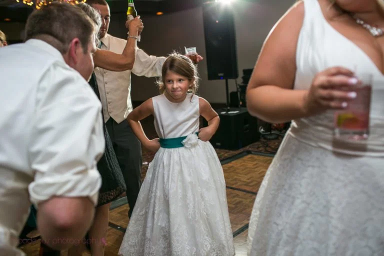 A young girl in a white dress with a teal sash stands with her hands on her hips, looking to the side during a social event, possibly a wedding reception, with adults around her holding drinks.