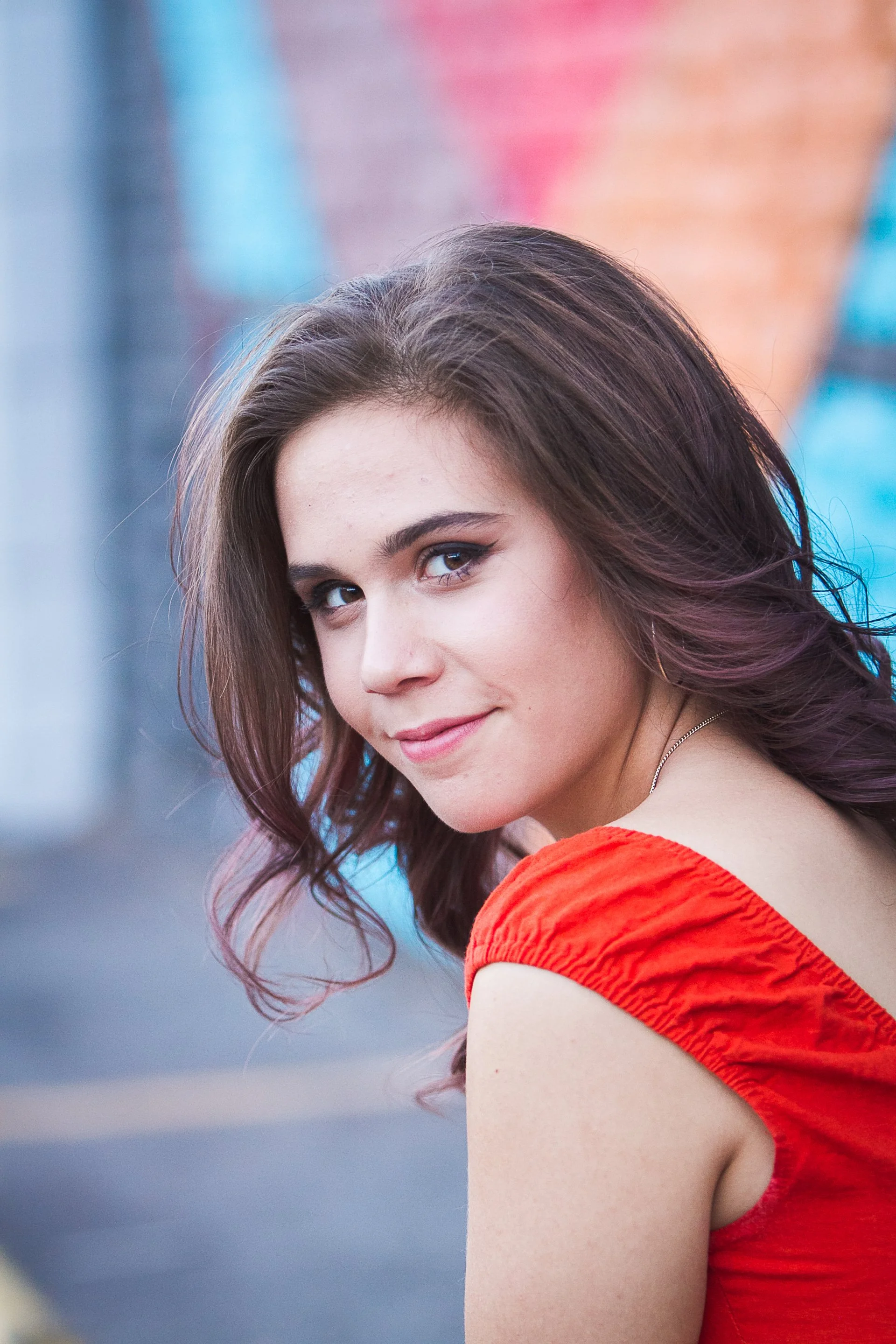 A young woman with wavy brown hair and blue eyes looks over her shoulder, wearing a red sleeveless top, with a colorful street wall in the background.