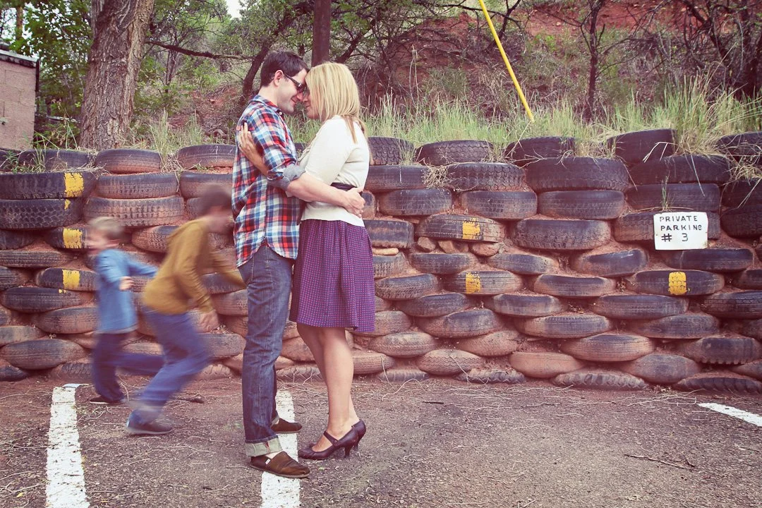 A young couple holding each other in front of a stack of tires, with two children playing in the background at a parking lot. There is a sign that reads 'PRIVATE PARKING #3'.