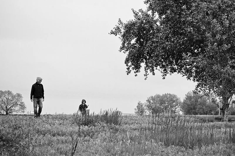 A man and a young girl walking outdoors in a grassy field with trees and overcast sky in the background.