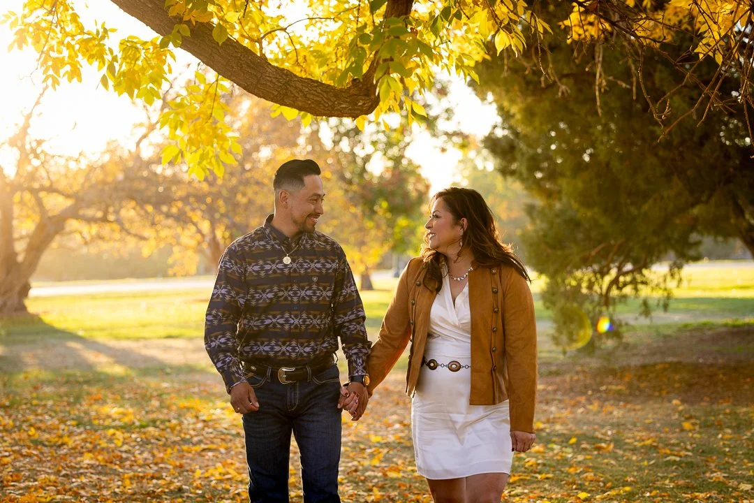 A couple holding hands and walking in a park during autumn, with trees and fallen leaves.