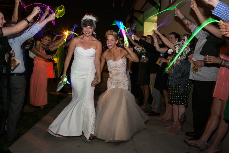 Two brides in wedding dresses walking down an aisle surrounded by friends and family holding glow sticks during a celebration.