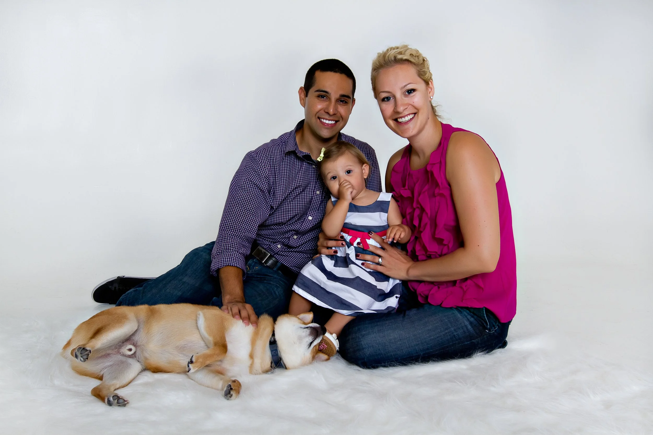 A happy family of three with a dog, sitting on a white furry rug against a plain white background. The father and mother are smiling, with the young daughter sitting between them, touching her nose with her hand. The small dog is lying on its side ne