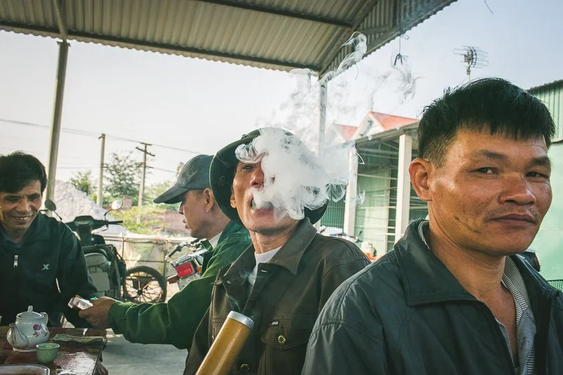 Group of men sitting together outdoors, with one man in the middle smoking and exhaling smoke, while others look on, in a casual setting.