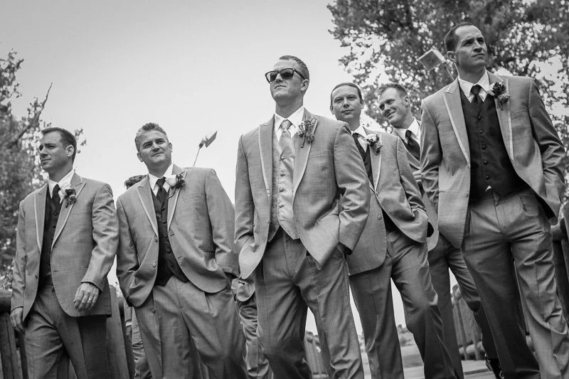 Group of six men dressed in suits standing outdoors, looking confident, with trees in the background.