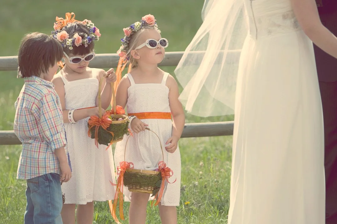 Three young girls in white dresses and flower crowns, wearing sunglasses, participate in a wedding ceremony outdoors. A woman in a wedding dress is visible on the right side of the image.