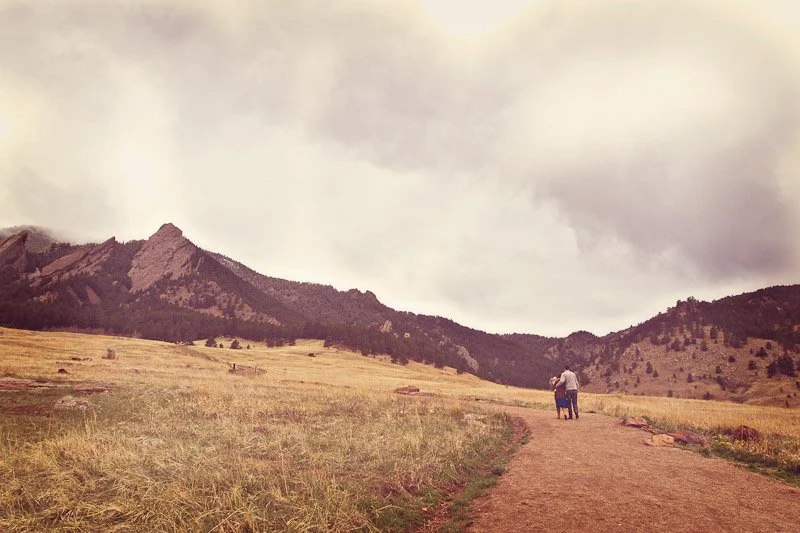 Two people walking on a dirt trail through a grassy field with mountains in the background under cloudy sky.