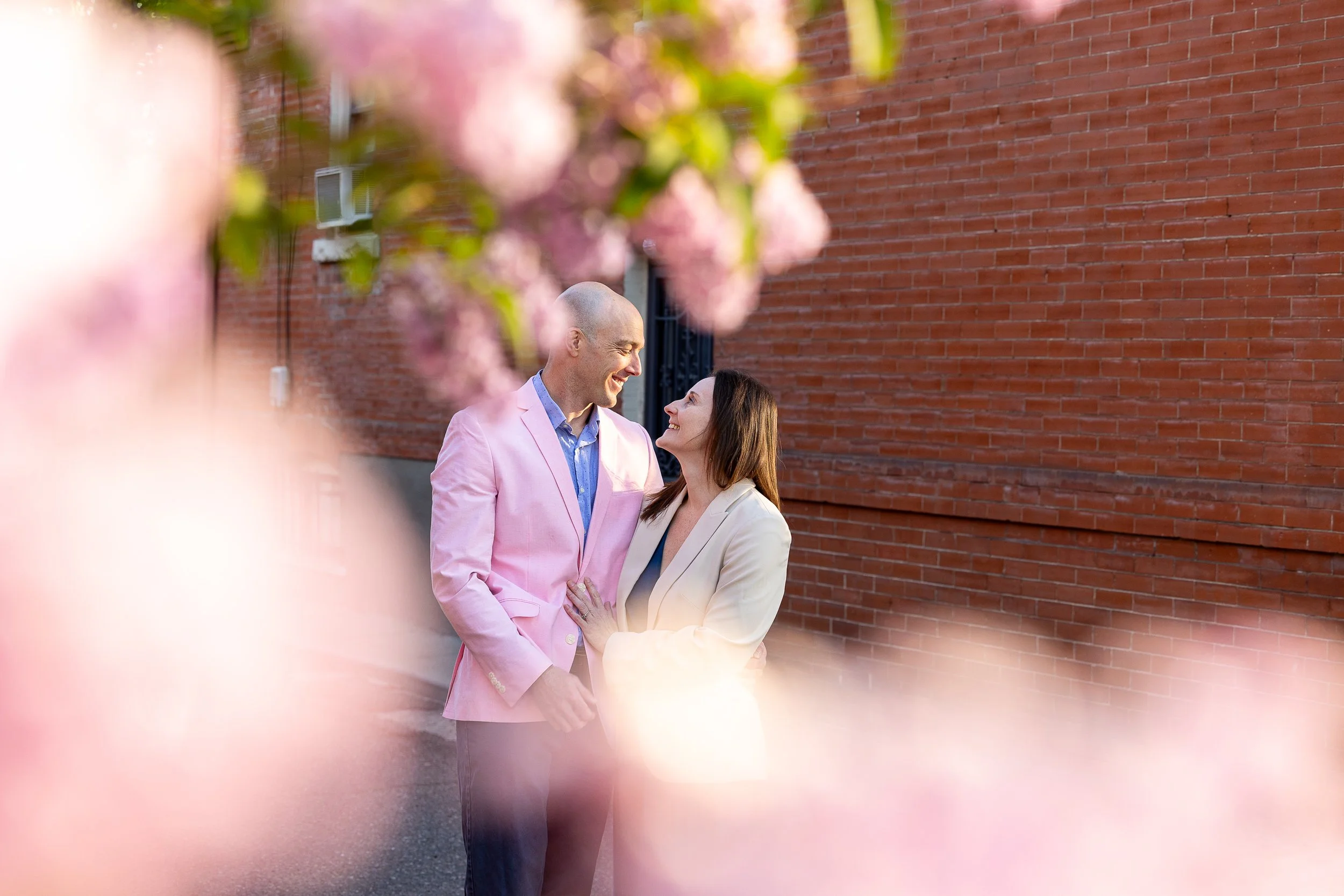 A couple standing close together outdoors, smiling at each other, framed by out-of-focus pink blossoms in the foreground, with a red brick wall in the background.