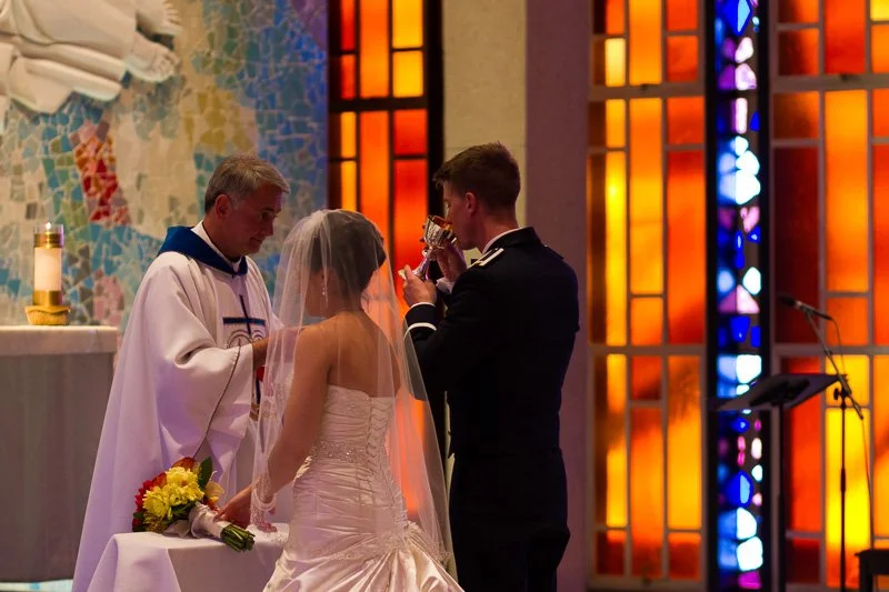 A bride and groom participate in a wedding ceremony in a church, with a priest present and colorful stained glass windows in the background.