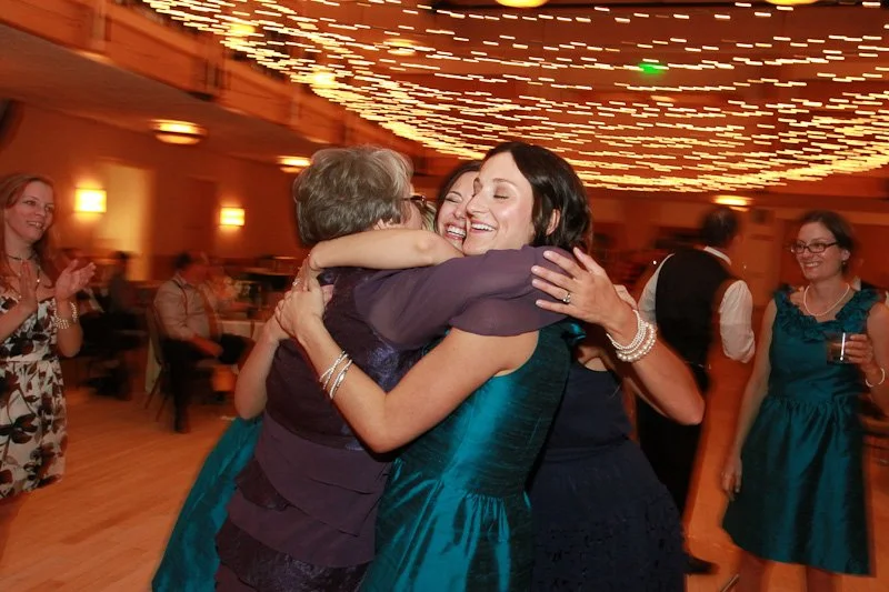People hugging and dancing at a celebration or wedding, with fairy lights on the ceiling in a banquet hall.