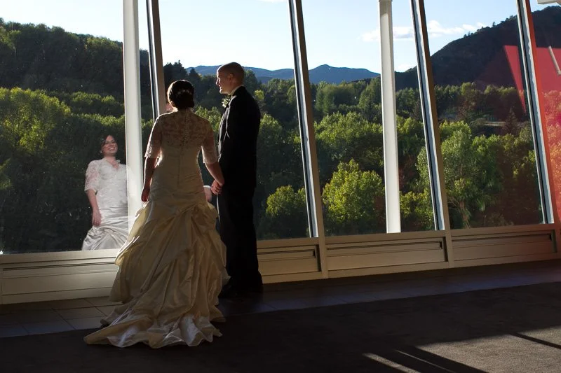 Bride and groom holding hands inside near large windows, with a woman outside in a white dress looking in, scenic mountain landscape in the background.