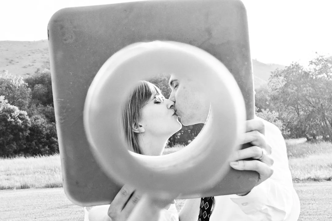 A man and woman kissing, viewed through a round mirror held by a person outside in a field with trees in the background.
