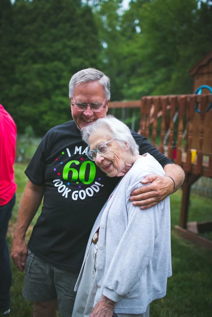 A man and an elderly woman sharing a hug outdoors in a green yard, with a wooden play structure in the background. The man is smiling and wearing a black t-shirt celebrating a 60th birthday.