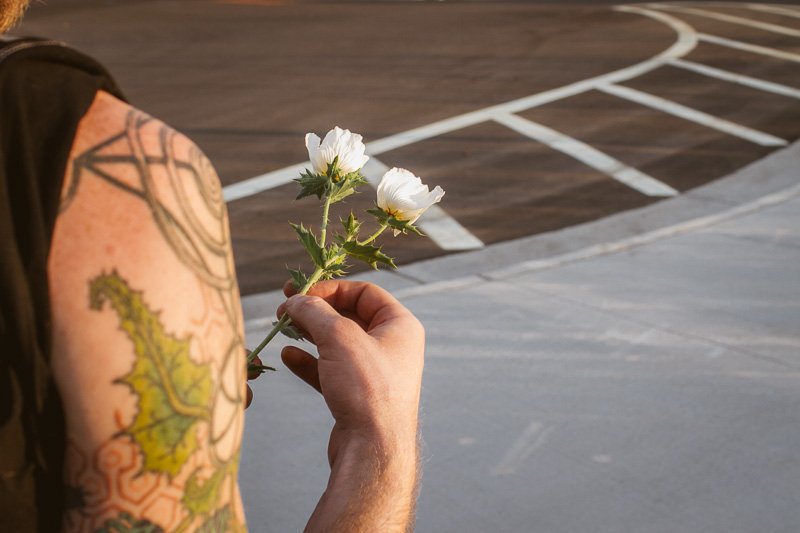A person with tattoos on their arm holding a white flower in front of their shoulder, with a parking lot background.