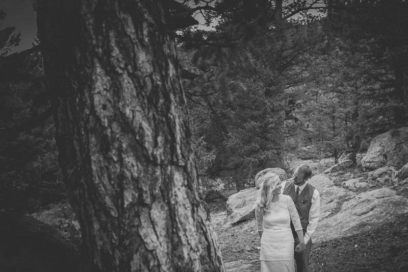 A couple holding hands in a forested outdoor setting, with large rocks and trees surrounding them.